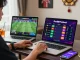 Engaged Thai bettor using Fun88Thailand on laptop and smartphone amidst lively atmosphere.